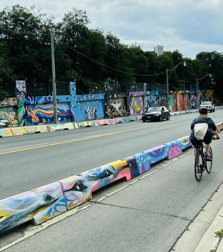 Photo of a Toronto highway-esque street during daylight (Lansdowne Ave). Two protected bike lanes on either side of the street are shown. There's a cyclist and 2 cars, all in motion and using their respective lanes, in the photo. There is a TON of colourful artwork in the photo that demands attention. It's on both sides (car and bike facing) of the half-metre tall bike lane barrier walls. It's also on the 3 or 4 metre high underpass-like walls of the street. About 150 metres of street artwork is capture in the photo