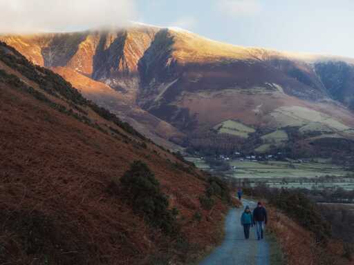 A landscape view from a hillside track in the Lake District looks across a valley towards the imposing, snow-dusted massif of Skiddaw. In the foreground, a man and a woman in winter coats and woolly hats walk towards the camera along a grey gravel path that winds around a steep slope covered in dry, rust-coloured bracken and dark green gorse. Dramatic golden sunlight breaks through the clouds, illuminating the upper ridges of the mountains in a warm glow while casting deep, cool shadows into the craggy ravines. Below the path, the valley floor is a patchwork of pale green fields and small clusters of white cottages, leading the eye towards the distant, hazy peaks under a soft, overcast sky.